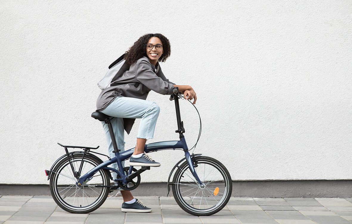 Junge Frau sitzt lachend auf einem Fahrrad mit grauen Jacke, Jeans und einem Rucksack. KARAWANE KiJU