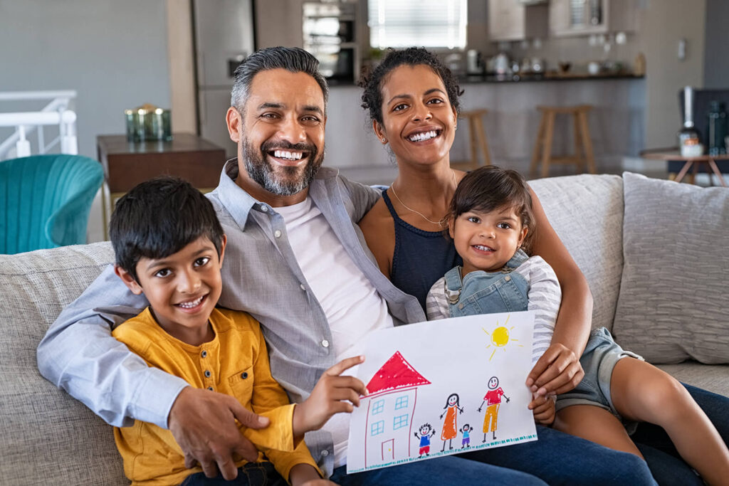 Eine Familie mit zwei Kindern sitzt lachend auf dem Sofa und halten ein selbstgemaltes Bild, welches eine Familie und ein Haus zeigt