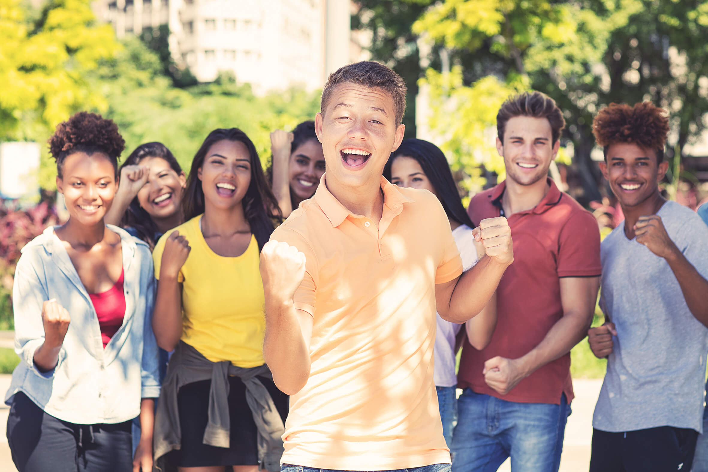 Ein Gruppe junger Menschen lachen in die Kamera. Im Vordergrund steht ein junger Mann mit orangenen Tshirt.